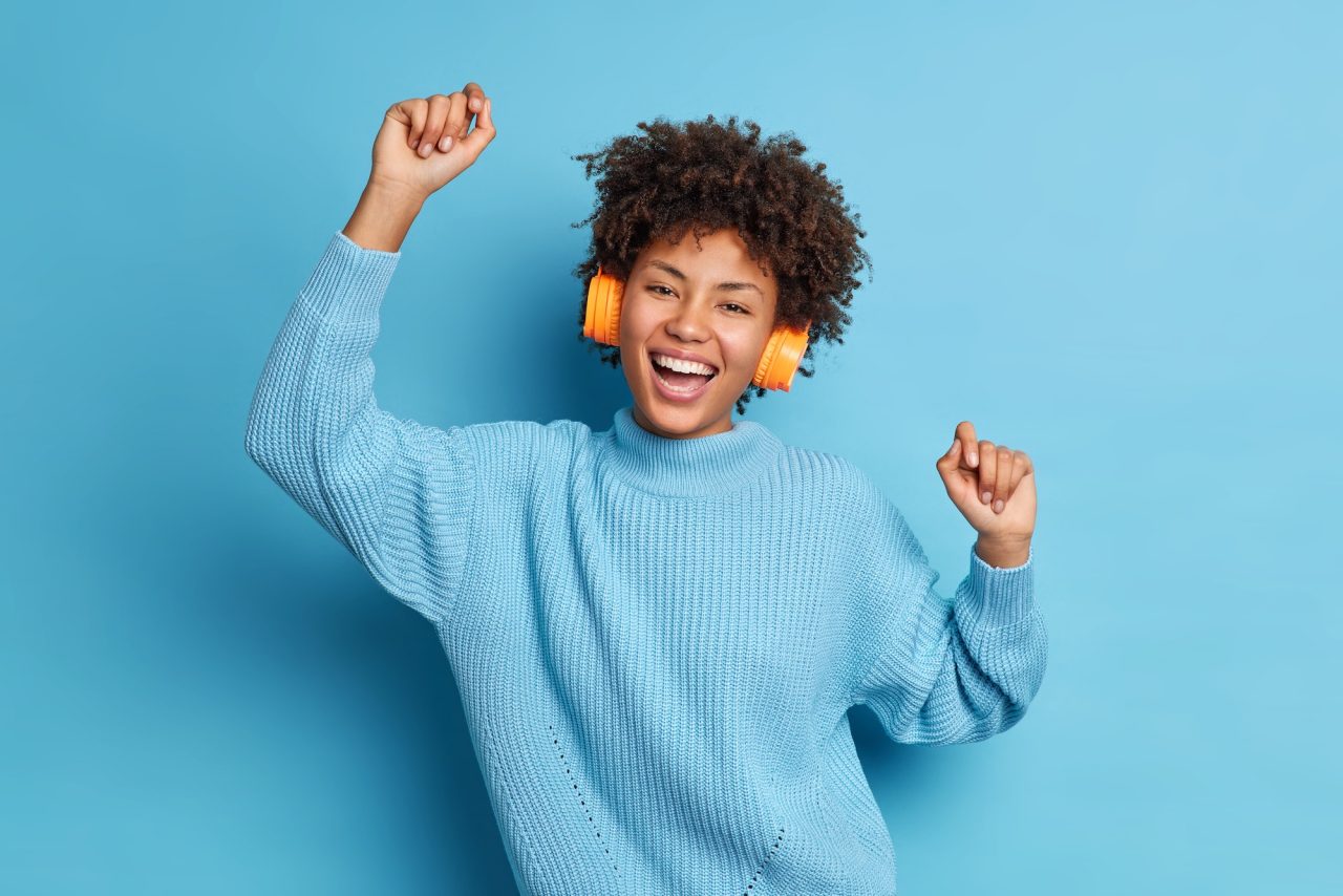 glad dark skinned woman with joyful expression raises arms and dances to music wears headphones and